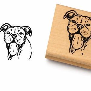May include: A black and white illustration of a dog's face, with its tongue hanging out, is on a wooden stamp. The stamp is rectangular and has a natural wood finish. The dog's features are detailed.