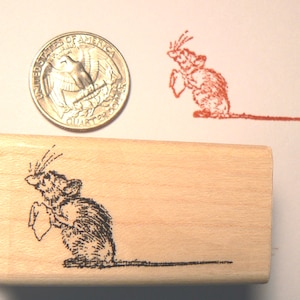 Pode incluir: Um carimbo de madeira com uma ilustração detalhada de um rato segurando um pequeno papel. Uma moeda americana está ao lado do carimbo para comparação de tamanho. Uma impressão em tinta vermelha do rato também é visível.