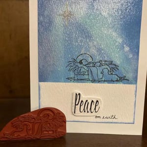 May include: A Christmas card with a blue watercolor background depicting a star and a nativity scene. The card has the text "Peace on earth" written on it.