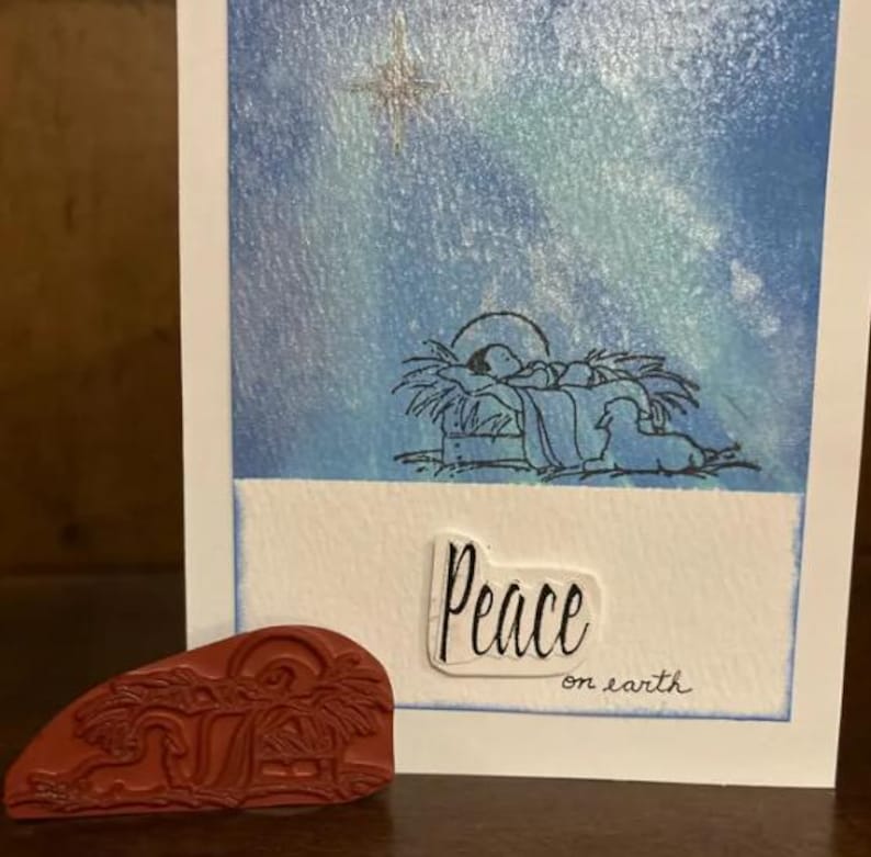 May include: A Christmas card with a blue watercolor background depicting a star and a nativity scene. The card has the text "Peace on earth" written on it.