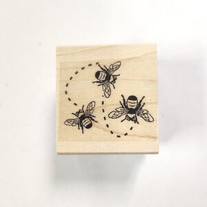 May include: A wooden stamp with a design of three bees flying in a circle. The bees are black and white and have a dotted line following their flight path.