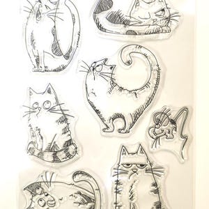 May include: A clear sheet of transparent stamps featuring various black and white illustrations of cats in different poses. The stamps are designed for crafting and scrapbooking.