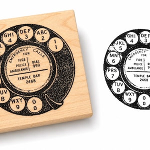 Puede incluir: Un sello de goma cuadrado de madera con un diseño en tinta negra de un dial de teléfono rotativo antiguo. El texto central dice EMERGENCY CALLS FOR FIRE POLICE AMBULANCE DIAL 999 y TEMPLE BAR 2468. El dial incluye los números del 1 al 0 y letras.