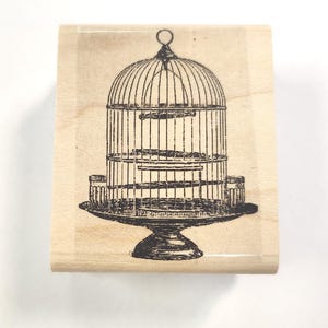 Könnte beinhalten: Ein Holz-Stempel mit einem Schwarzweißbild eines Vogelkäfigs auf einem Sockel. Der Vogelkäfig hat ein gewölbtes Dach und drei Ebenen.