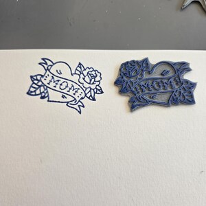 May include: Two blue ink stamps on white paper. One stamp is a heart with a banner that reads "MOM" and a rose. The other stamp is a similar design, but with a different shape. The stamps are likely used for crafting or scrapbooking.
