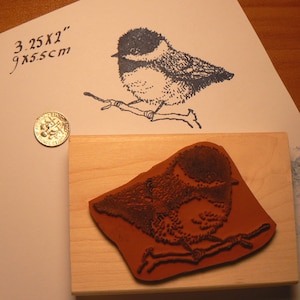 Puede incluir: Un sello de goma con una base de madera marrón. El sello presenta una imagen detallada de un pájaro posado en una rama. La imagen mide 3,25 x 2 pulgadas o 9 x 5,5 cm.