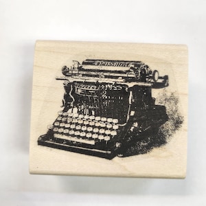 May include: A vintage typewriter rubber stamp with a black and white image of a typewriter. The stamp is made of wood and has a rectangular shape.