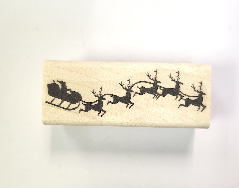 May include: A wooden stamp with a black silhouette of Santa Claus in a sleigh pulled by reindeer. The reindeer are in a line, pulling the sleigh with a rope.