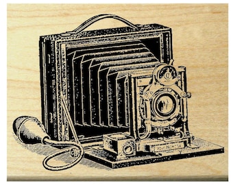 May include: A black and white illustration of a vintage camera with a bellows design, lens, and handle. The camera is on a wooden surface. The illustration includes a bulb and a small box.