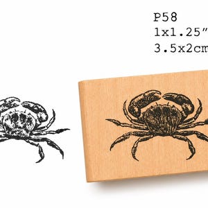 Puede incluir: Un sello de madera con una detallada ilustración de un cangrejo negro. El sello mide 3,5x2cm. La imagen también muestra una ilustración de cangrejo más pequeña y separada. El texto "P58" está impreso encima del sello.