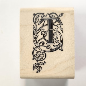Könnte beinhalten: Ein Stempel aus Holz mit einem schwarzen Tintenmuster eines dekorativen Buchstabens T. Der Buchstabe ist von einem floralen Muster umgeben.