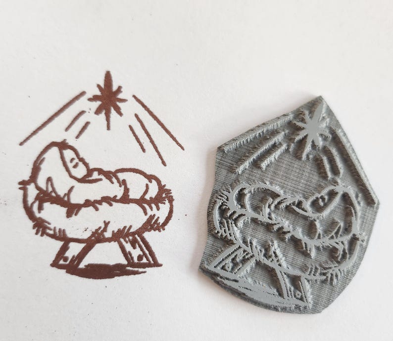 May include: A gray rubber stamp with a raised design of a baby in a manger with a star above. The stamp is designed to be used for creating a Christmas card or other holiday craft.