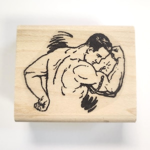May include: A black and white illustration of a muscular man flexing his bicep. The image is printed on a wooden block.