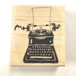May include: A wooden stamp featuring a black illustration of a vintage typewriter with a sheet of paper inserted. The stamp includes a stylized signature in black ink. The wooden block has a light wood grain.