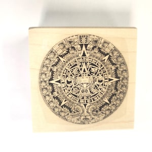 Puede incluir: Un sello de madera con una imagen en blanco y negro del calendario azteca, también conocido como la Piedra del Sol, sobre un fondo marrón claro.