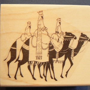 May include: A woodblock stamp with a black and white illustration of three figures riding camels. The figures are wearing robes and crowns. The camels are walking in a line.