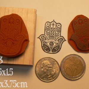 Pu&ograve; includere: Due timbri in gomma arancione con un design a mano di Hamsa, uno montato su un blocco di legno, accanto a un'impronta a inchiostro nero. Una moneta da 25 centesimi USA e una da 2 euro indicano la scala, con il testo P63, 1.25x1.5, 3.5x3.75cm.