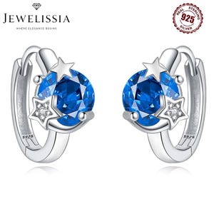 May include: Silver hoop earrings with a star and blue gemstone design. Each earring has a large, faceted blue gemstone, a small star, and a star-shaped accent with a small diamond. The earrings are marked with "S925".