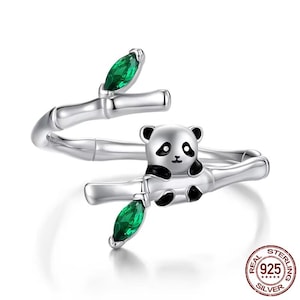 May include: A silver ring with a panda bear charm sitting on a bamboo branch with green crystal accents. The ring is marked "REAL STERLING 925 SILVER".