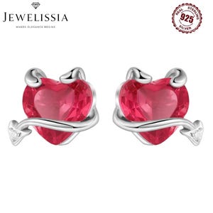 Può includere: Un paio di orecchini a lobo a forma di cuore. Ogni orecchino presenta un cuore rosso vibrante abbracciato da un design in metallo color argento. Piccole gemme trasparenti sono incastonate alle estremità degli accenti argentati. Gli orecchini sono su sfondo bianco.