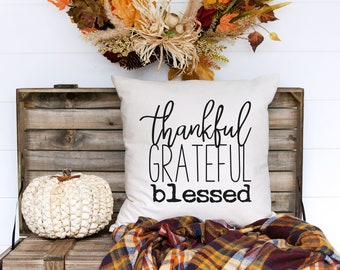 May include: A white throw pillow with black text that reads "thankful grateful blessed". The pillow is sitting on a wooden crate with a white pumpkin and a plaid blanket.