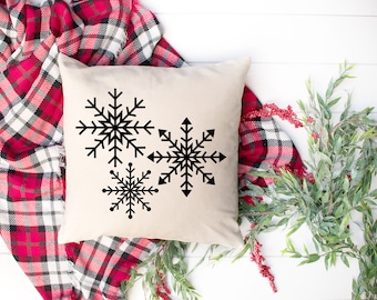 May include: A white throw pillow with three black snowflake designs. The pillow is on a red and white plaid blanket.