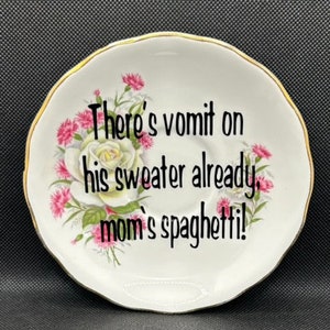 May include: White ceramic saucer with a gold rim and floral design. The saucer has a black text message that reads "There's vomit on his sweater already, mom's spaghetti!"