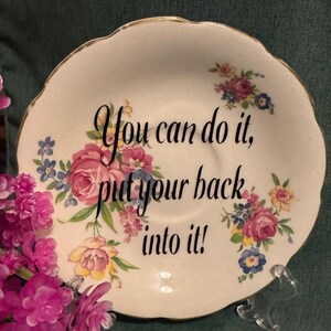 May include: A white porcelain saucer with a gold rim and floral pattern. The saucer has the text "You can do it, put your back into it!" printed on it.