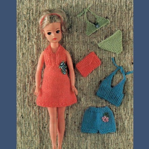 May include: A doll wearing a knitted coral dress with a floral design. The doll is surrounded by knitted clothing items, including a green bikini, a blue bikini, and a coral purse.