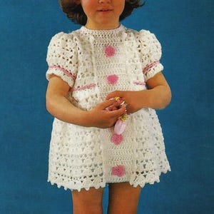 May include: A white crochet dress with pink detailing and buttons. The dress has short, puffed sleeves and a scalloped hem. The dress is worn by a young child against a blue backdrop.