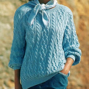 May include: A light blue cable-knit sweater with three-quarter sleeves. The sweater has a round neckline and a textured pattern. A light blue scarf is tied around the neck. The person is wearing blue jeans.