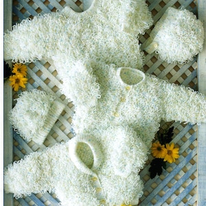 Könnte beinhalten: Eine Sammlung von Babykleidung aus weichem, strukturiertem Strick. Das Set beinhaltet zwei Strickjacken, einen Pullover und zwei passende Mützen. Die Kleidungsstücke sind cremefarben mit blauen und gelben Tupfen und werden auf einem hellblauen Gitterhintergrund mit gelben Blumen präsentiert.