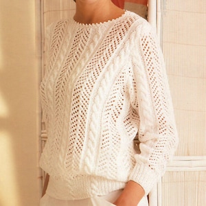 May include: A white knitted sweater with a round neckline and long sleeves. The sweater features a textured pattern with openwork details and cable knit accents. The bottom hem and cuffs are ribbed.