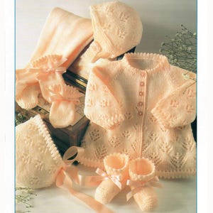 May include: A collection of soft peach-colored knitted baby clothes, including a jacket, bonnet, booties, mittens, and a blanket. The jacket has button closures and a delicate leaf pattern. The booties and mittens are adorned with satin bows.