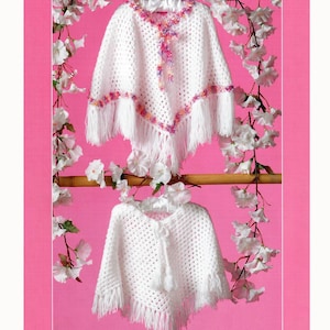 May include: Two white crochet ponchos with fringe and floral accents. The top poncho has pink and purple trim and a decorative closure. The bottom poncho has a tassel closure. Both are displayed against a pink background with white flowers.
