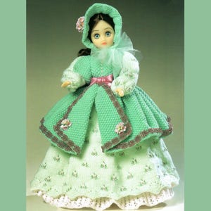 May include: A knitted doll wearing a green dress with a white lace trim and a green hooded coat. The doll has brown hair and blue eyes. The dress is decorated with small white flowers.