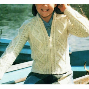 May include: Cream-colored, cable-knit cardigan with a full zipper. The sweater features a textured diamond and cable pattern. The person is wearing a blue collared shirt underneath. The background shows a body of water and a boat.