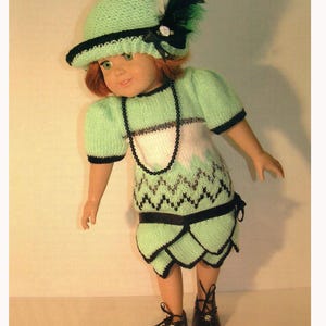 Puede incluir: Una muñeca vestida con un conjunto de punto verde claro con ribetes negros, que incluye un sombrero con una pluma y un vestido a juego. El vestido tiene un patrón en zigzag blanco y negro y un cinturón negro. La muñeca también lleva zapatos negros y un collar de cuentas.