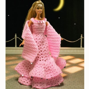 Op de afbeelding: Een roze gehaakte poppenjurk met lange mouwen en een gelaagde rok. De jurk is versierd met kleine, zilveren pailletten en wordt gedragen door een blonde pop. De achtergrond toont een nachtelijke hemel met een maan en sterren.