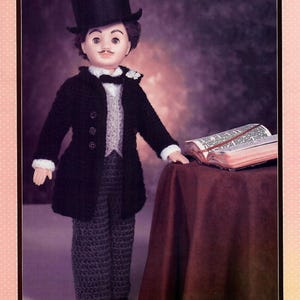 May include: A crochet doll dressed in a black top hat, coat, and pants, with a white vest and bow tie. The doll has a painted face with a mustache and is standing next to a table with a book.