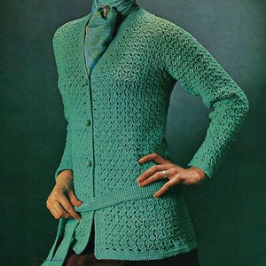 May include: A seafoam green crochet cardigan with long sleeves and a button-up front. The cardigan features a textured stitch pattern and a matching belt. The model is wearing a scarf with a brooch.