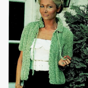 May include: A light green, short-sleeved jacket with a collar. The jacket is made of a textured, fuzzy material. The model is wearing a white top and a white belt. The background includes a green bush and a white wall.
