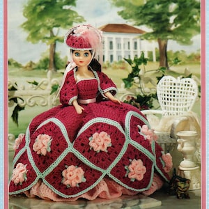 Pode incluir: Boneca com um vestido e chapéu de crochê bordô, com detalhes em rosa e azul claro. O vestido apresenta um padrão quadrado com detalhes florais rosa e bordas azuis claras. A boneca está posada em frente a uma cena de jardim.