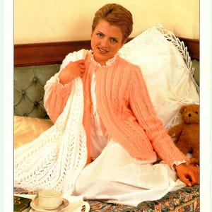 Könnte beinhalten: Eine Frau trägt einen pfirsichfarbenen Strickcardigan mit einem Zopfmuster. Sie sitzt auf einem Bett mit einer weißen Strickdecke und einem Teddybär.