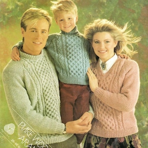 Puede incluir: Una familia de tres personas con suéteres de punto de cable a juego. Los suéteres son en tonos de verde claro, azul y rosa. Los suéteres presentan un patrón de punto de cable texturizado. El texto "PURE WOOL ARAN" es visible en la esquina inferior izquierda de la imagen.