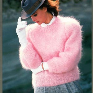 Op de afbeelding: Een lichtroze, pluizige gebreide trui met lange mouwen. De trui wordt gecombineerd met een grijze hoed met een bruine band, witte handschoenen en een witte overhemdkraag. Het model draagt een grijs-witte geruite rok.