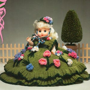 May include: A vintage doll with blonde hair and blue eyes, wearing a green knitted dress adorned with pink, blue, and white floral accents. The doll has a matching hat and a ribbon around its neck. A small topiary and a white picket fence are in the background.