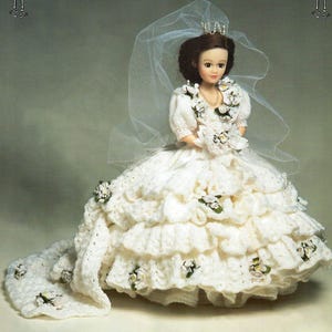 Puede incluir: Muñeca vestida con un vestido de novia blanco de ganchillo hecho a mano con una larga cola. La muñeca tiene cabello castaño, una tiara y un velo. El vestido está adornado con pequeñas flores blancas y hojas verdes.