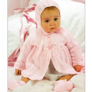 May include: A pink knitted baby outfit with a matching hat and booties. The outfit features a delicate lace pattern and a bow at the neckline.