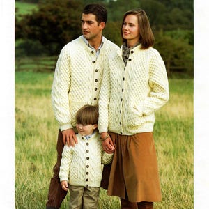 May include: A family of three wearing white cable knit cardigans. The adults are standing and holding hands with a young child. The child is wearing a white cable knit cardigan and khaki pants.
